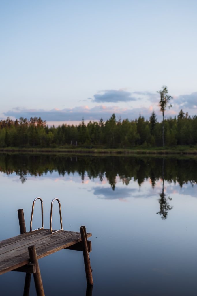 a wooden jetty over a lake that is so still that it has a perfect reflection of the trees and the pink skies in the water during sunset in sweden